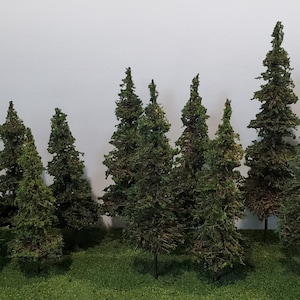 May include: A collection of eleven miniature model pine trees with textured green and brown foliage, arranged in a cluster on a flat, rectangular green base that mimics a grassy surface, creating a dense forest scene for hobby dioramas.