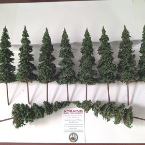 May include: A collection of model trees and bushes for miniature landscapes. The trees are green with brown stems, and the bushes are a darker green. The trees are approximately 6 inches tall. The items are arranged on a white surface.