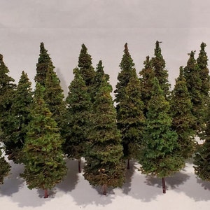 May include: A collection of 14 miniature evergreen trees, each with a brown trunk and green foliage. The trees are arranged in a row on a white surface.