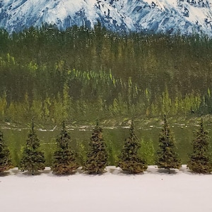 Puede incluir: Una fila de 12 árboles de hoja perenne en miniatura, pintados en tonos de verde, están dispuestos sobre una superficie blanca frente a una pintura de una cordillera nevada y un bosque. Los árboles miden aproximadamente 2,5 cm de alto.