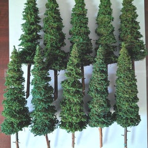 May include: Nine miniature evergreen trees with green foliage and brown trunks are arranged on a white surface. The trees vary in height and are designed for model landscapes or craft projects.