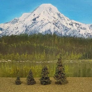 May include: A painting of a snow-capped mountain peak rising above a dense green forest and a calm lake. In the foreground, four small, three-dimensional model evergreen trees of varying heights are arranged in a row on a textured brown surface.