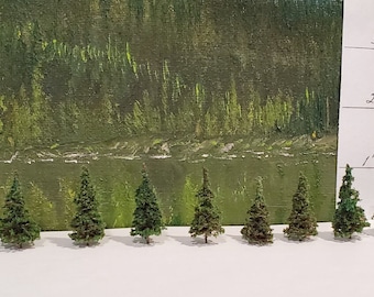Op de afbeelding: Tien miniatuur-naaldbomen, variërend in grootte van 2,5 tot 7,5 cm hoog, zijn in een rij op een wit oppervlak gerangschikt. Een liniaal met duimmarkeringen is zichtbaar op de achtergrond.