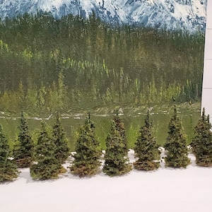 May include: A collection of 17 miniature evergreen trees, ranging in size from 1 to 4 inches tall, arranged in a row on a white surface. The trees are made of green and brown materials, resembling a miniature forest.