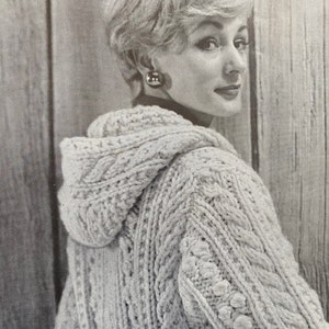 May include: A close-up of a woman wearing a white knitted sweater with a hood and cable knit detail. The sweater has a textured pattern and a loose fit.