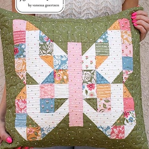 May include: A quilted butterfly pillow with a green background and a patchwork design in white, pink, orange, and blue. The pillow has a stitched border and the text "Lella Boutique" and "Mini Charmed".