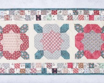 May include: A white table runner with a teal border and three red, white, and blue floral designs. The floral designs are made of patchwork fabric squares.