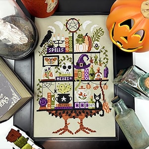 May include: Cross-stitch artwork with a Halloween theme, featuring the words "Spells", "Hexes", and "Potions". The design includes a black cat, a skull, a witch's hat, and potion bottles. The artwork is framed in black.