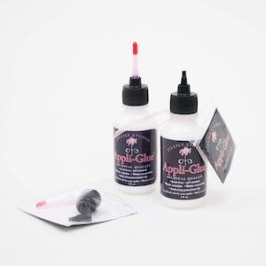 May include: Two white bottles of Appli-Glue archival quality adhesive with black caps and red applicator tips. A small bag contains extra applicator parts. The bottles have labels with the text "Appli-Glue" and "JILLILY STUDIO".