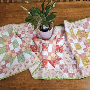 May include: A rectangular quilted table runner featuring a patchwork star pattern in shades of green, yellow, and red. The fabric has a floral print border and a light green edge binding. A small potted green plant sits in the center of the runner.
