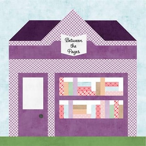 May include: A fabric quilt block depicting a purple storefront with a white sign reading Between the Pages. The building features a purple roof, a white door, and two display windows filled with rows of colorful patterned fabric books.