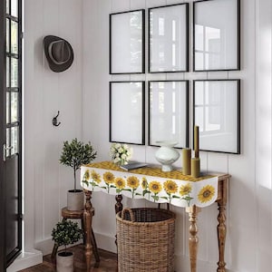 May include: A hallway scene featuring a wooden table with a sunflower-patterned runner, a woven basket, and framed artwork. The table is adorned with a vase, candles, and a small plant. A hat hangs on the wall, and a small stool holds another potted plant.