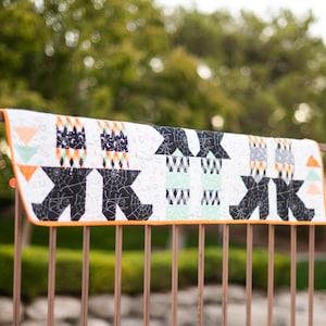 May include: A Halloween-themed quilt with a white background, featuring black spiderwebs, orange triangles, and geometric patterns. The quilt is trimmed with orange binding and displayed on a brown metal fence.
