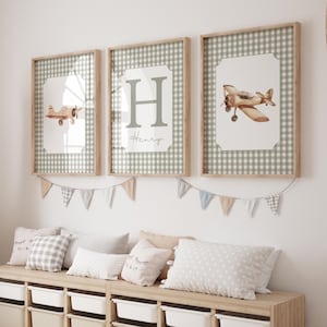 May include: Three framed wall art pieces with light brown wooden frames. The art features a watercolor airplane, the letter "H" with the name "Henry", and a gingham pattern. A garland of small flags hangs below the art, and pillows are on a wooden bench.