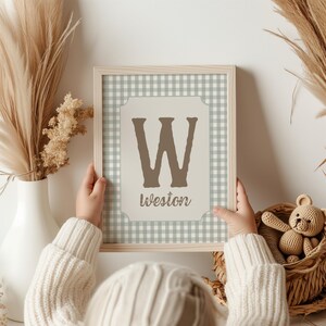 May include: A framed print featuring a large brown letter "W" and the name "Weston" in a decorative font. The background has a light green and white gingham pattern. The frame is light wood. The print is held by a person wearing a white sweater.