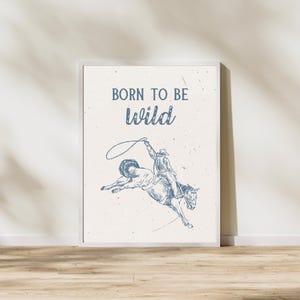 K&ouml;nnte beinhalten: Ein wei&szlig;er gerahmter Druck mit dem Text "Born to be wild" in blauer Schrift. Unter dem Text befindet sich eine blaue Strichzeichnung eines Cowboys, der ein buckelndes Pferd reitet.