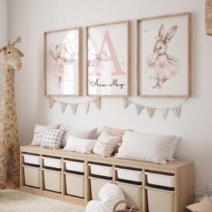 May include: Three framed prints with a pink and white color scheme. The prints feature a ballerina bear, a ballerina bunny, and a pink letter A with the name "Ava May" written below. The prints are hanging on a white wall above a wooden bench with storage bins.