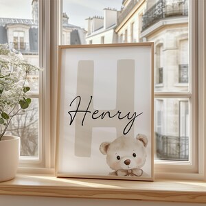 Puede incluir: Un cuadro enmarcado con el nombre "Henry" en escritura cursiva, sobre una gran "H". Una ilustraci&oacute;n de un oso de peluche en la parte inferior. La obra de arte se exhibe en un alf&eacute;izar de madera, con vistas a edificios.