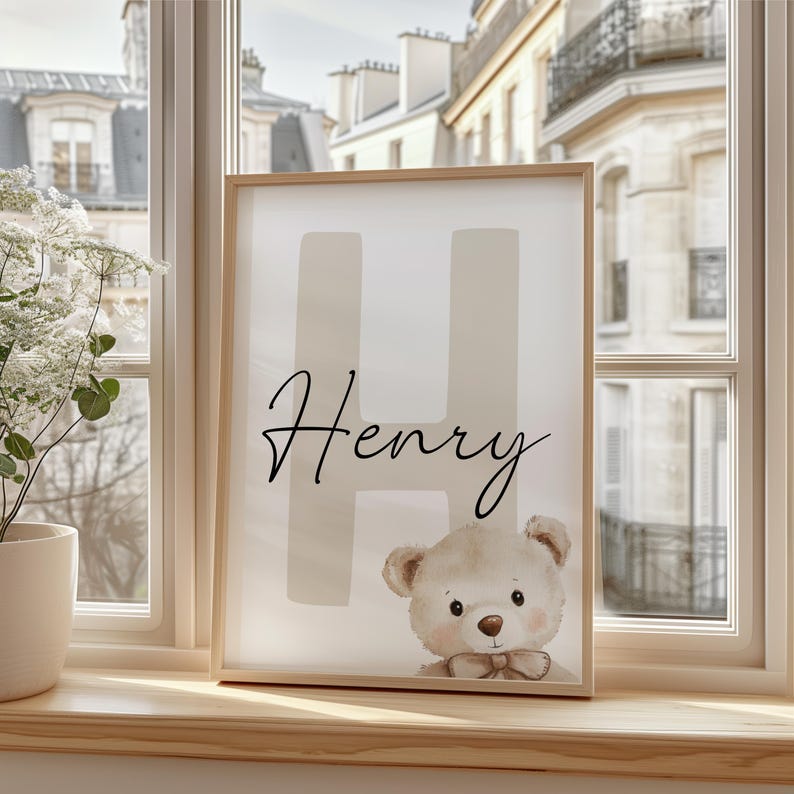Puede incluir: Un cuadro enmarcado con el nombre "Henry" en escritura cursiva, sobre una gran "H". Una ilustraci&oacute;n de un oso de peluche en la parte inferior. La obra de arte se exhibe en un alf&eacute;izar de madera, con vistas a edificios.