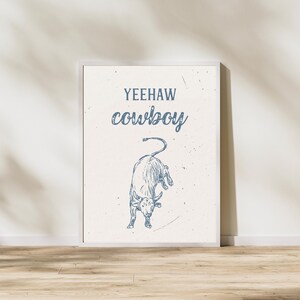K&ouml;nnte beinhalten: Ein wei&szlig;er gerahmter Druck mit einer blauen Bullenillustration und dem Text "Yeehaw cowboy" in Blau.