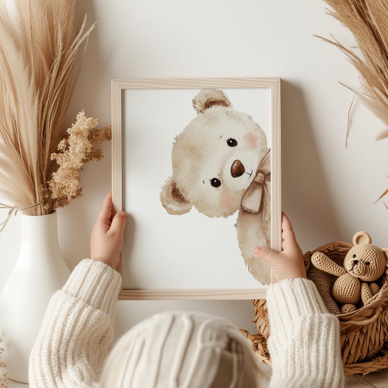 Puede incluir: Una impresi&oacute;n enmarcada con una ilustraci&oacute;n en acuarela de un oso de peluche asom&aacute;ndose por la esquina. El oso tiene una nariz marr&oacute;n, un lazo y mejillas rosadas. El marco es de madera clara. Un peque&ntilde;o oso de peluche de ganchillo est&aacute; en una cesta.