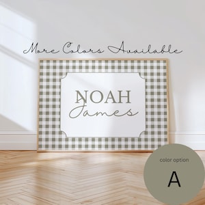 May include: A framed wall art piece featuring the name "Noah James" in gray script and block letters. The background has a gray and white gingham pattern. The text "More Colors Available" is written above the frame. A color option A is shown in a circle.