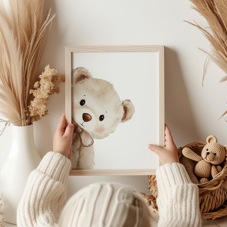 Puede incluir: Una impresi&oacute;n enmarcada con una ilustraci&oacute;n de acuarela de un oso de peluche mirando de reojo. El oso tiene pelaje marr&oacute;n claro, una nariz marr&oacute;n y mejillas rosadas. El marco es de madera clara. Un osito de peluche tejido est&aacute; en una cesta.