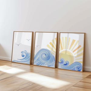 May include: Three framed watercolor paintings depicting a stylized ocean scene with blue waves and a yellow sun. The first painting shows a wave with a few white birds flying above. The second painting shows a wave with a sun rising behind it. The third painting shows a wave with a sun rising behind it.