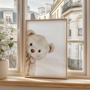 Puede incluir: Una impresi&oacute;n enmarcada con una ilustraci&oacute;n de acuarela de un oso de peluche asom&aacute;ndose. El oso tiene pelaje marr&oacute;n claro, una nariz marr&oacute;n y un peque&ntilde;o lazo. El marco es de madera clara.
