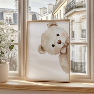 Puede incluir: Impresi&oacute;n enmarcada con una ilustraci&oacute;n de acuarela de un oso de peluche mirando de reojo. El oso es de color marr&oacute;n claro con un lazo a juego y mejillas rosadas. El marco es de madera clara y la impresi&oacute;n est&aacute; en un alf&eacute;izar.