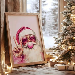 May include: A framed watercolor painting of Santa Claus wearing pink sunglasses and blowing a pink bubble. He is giving the peace sign with his right hand. The background is a light pink color.