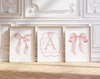 May include: Three framed prints with a light pink bow design. The center print features the letter "A" and the name "Annabeth". The prints are displayed on a wooden floor against a white wall.