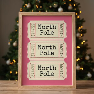 May include: Three vintage-style tickets with the text "North Pole" printed on them. The tickets are framed in a light wood frame and have a pink background.