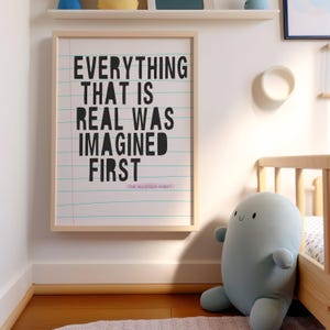 May include: A framed print with the quote "Everything that is real was imagined first" by The Velveteen Rabbit on a white background with pink lines. The print is hanging on a white wall next to a light blue plush toy.