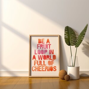 Puede incluir: Una impresión enmarcada con una cita motivacional en estilo acuarela. La cita dice "Sé un fruit loop en un mundo lleno de cheerios". La impresión está colgada en una pared blanca con una planta en maceta y esferas decorativas en primer plano.