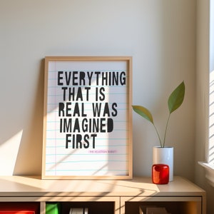 May include: A framed print with a white background and black text that reads "Everything that is real was imagined first." The text is on a blue and pink lined background. The bottom of the print has the text "The Velveteen Rabbit" in smaller font. The print is sitting on a light brown wooden shelf.