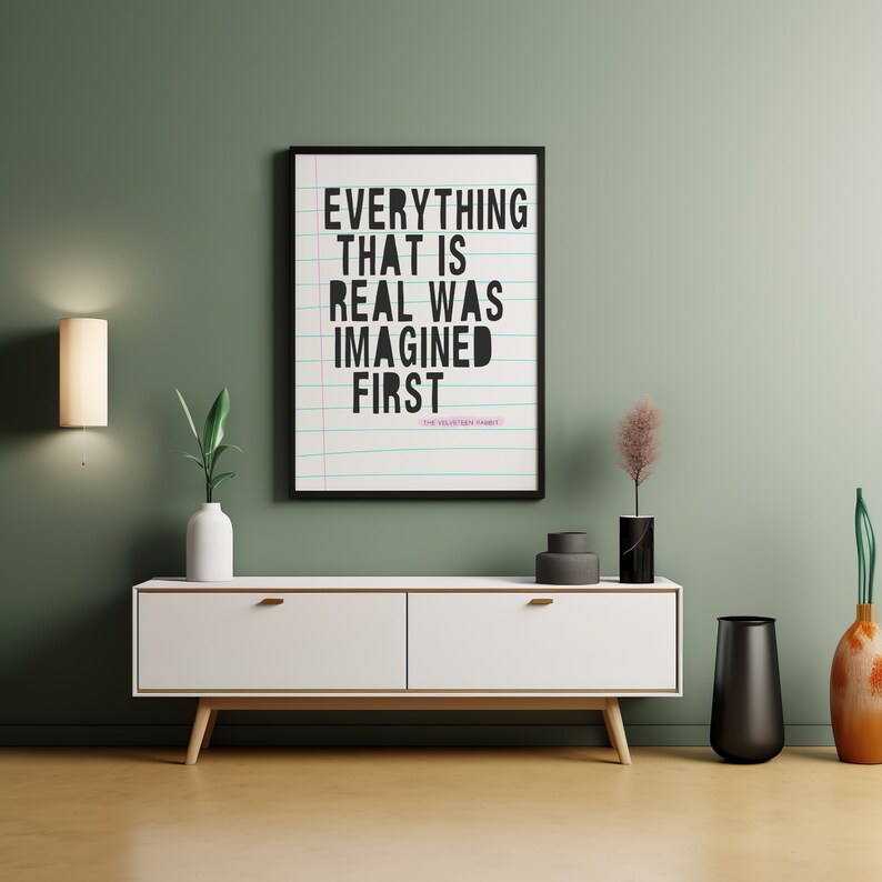 May include: A white media console with two drawers and wooden legs. A black and white framed print with the text "EVERYTHING THAT IS REAL WAS IMAGINED FIRST - THE SELECTION" hangs on a sage green wall above the console. A vase with dried flowers, a black vase, and a vase with orange flowers are on the console.