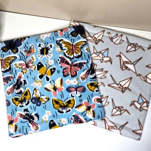 May include: Two square fabric pot holders. One features a light blue background with a colorful pattern of various butterflies in shades of yellow, pink, black, and white. The other has a light gray background with a pattern of brown-outlined origami cranes.