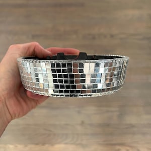 Puede incluir: Un cenicero redondo plateado con diseño de bola de discoteca, con interior negro y una muesca para apoyar objetos. El cenicero está cubierto de pequeñas baldosas cuadradas reflectantes, creando un efecto brillante. El cenicero está sostenido en una mano.