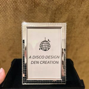 Może przedstawiać: Dekoracyjna ramka na zdjęcia z lustrzanym wzorem mozaiki. Ramka zawiera białą kartę z grafiką kuli dyskotekowej i tekstem "A DISCO DESIGN DEN CREATION". Ramka znajduje się na czarnej powierzchni.