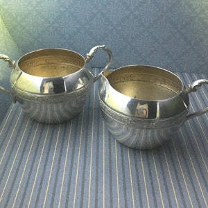 Pode incluir: Duas peças de servir prateadas, um cremeira e um açucareiro, estão lado a lado. Ambas têm alças ornamentadas e uma faixa decorativa. Os interiores têm um acabamento dourado. As peças estão sobre uma superfície listrada azul e branca.