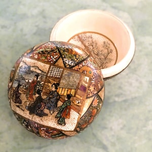 May include: A round, ceramic trinket box with a lid. The box is decorated with a colourful Japanese scene of people in traditional clothing. The lid is plain white on the inside.