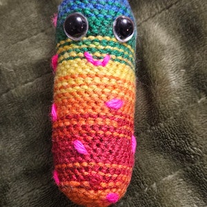 May include: A rainbow-colored crocheted worm with pink accents and black button eyes. The worm is smiling.