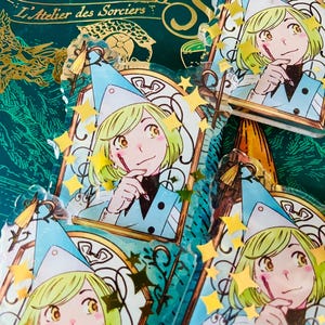 May include: Several clear acrylic charms featuring a cartoon character with green hair, a blue hat, and a blue outfit. The charms are decorated with gold stars and a gold frame. The background includes text that reads "Art des Sorciers".