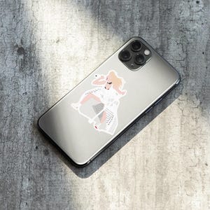 May include: A silver iPhone with a white sticker of a woman in a white dress and blonde hair. The sticker is on the back of the phone.