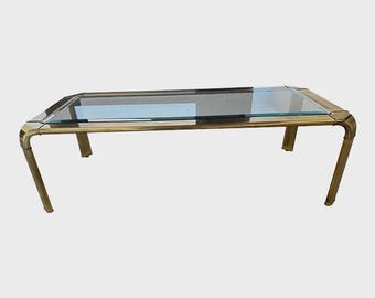 May include: Rectangular coffee table with a clear glass top and a brass-colored metal frame. The table has four legs and rounded corners. The glass top is approximately 48 inches long and 24 inches wide.