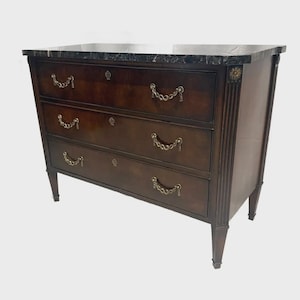 May include: A dark brown wooden dresser with three drawers and a black marble top. The drawers have gold-colored chain pulls and keyhole details. The dresser has a classic design with decorative side panels and tapered legs.