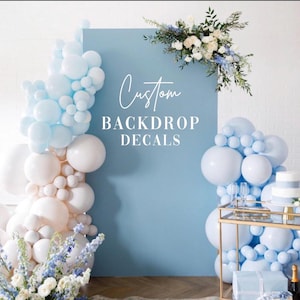 Puede incluir: Un telón de fondo azul claro con las palabras "Custom BACKDROP DECALS" en escritura blanca. El telón de fondo está decorado con globos blancos y azul claro, y arreglos florales. Un carrito de bar dorado está en primer plano.