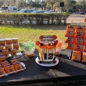 Op de afbeelding: Een verjaardagstafel met basketbalthema. De tafel heeft een versierde taart met oranje glazuur, een "Happy Birthday" bord en het nummer 12. Er zijn ook oranje traktatiezakjes, bekers en snoep.