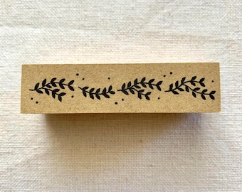 May include: A rectangular, wooden stamp with a decorative black leaf and dot border design. The stamp is a light brown color and is set against a white fabric background. The leaves are stylized and arranged in a repeating pattern.
